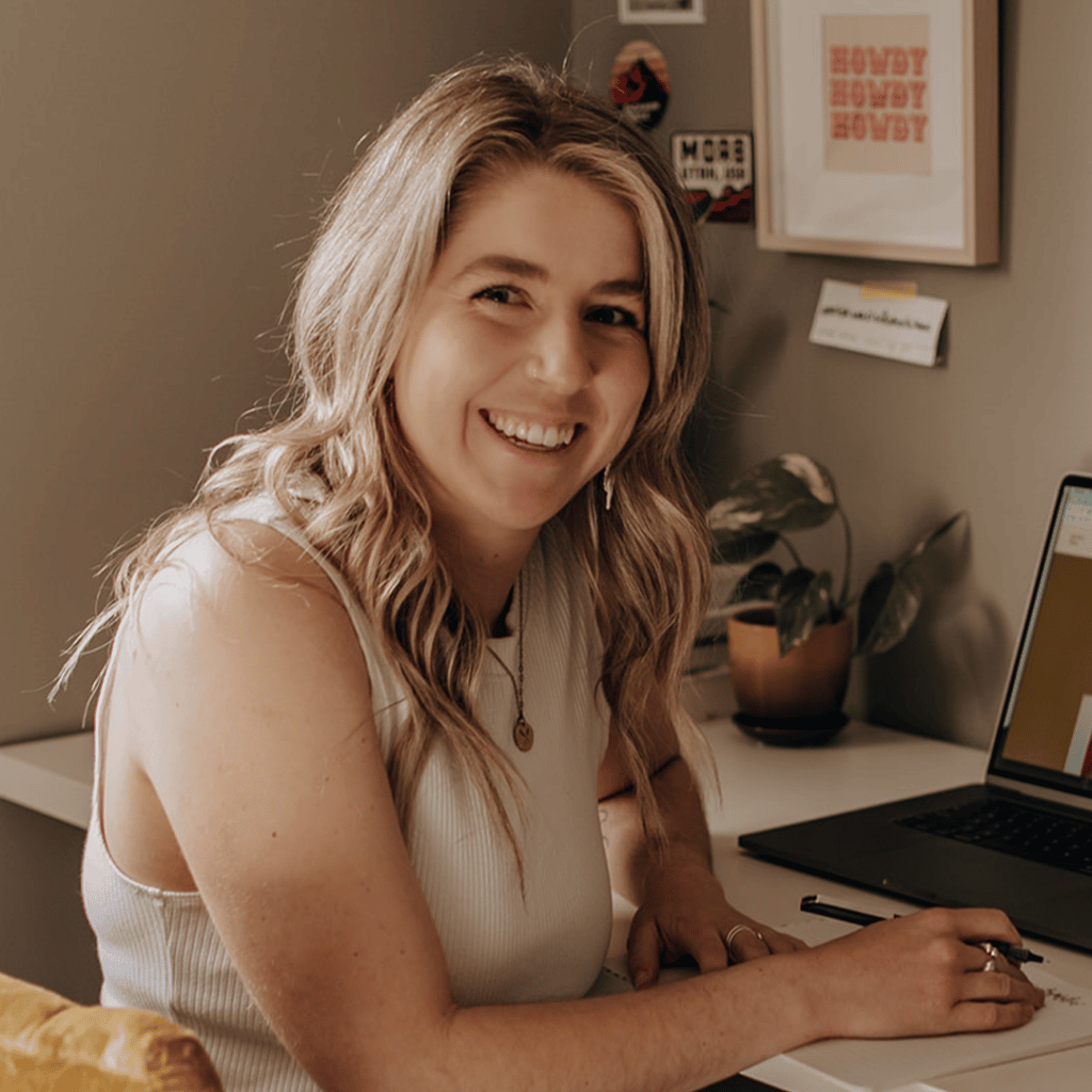Smiling woman with long blonde hair sitting in front of a laptop holding a pen