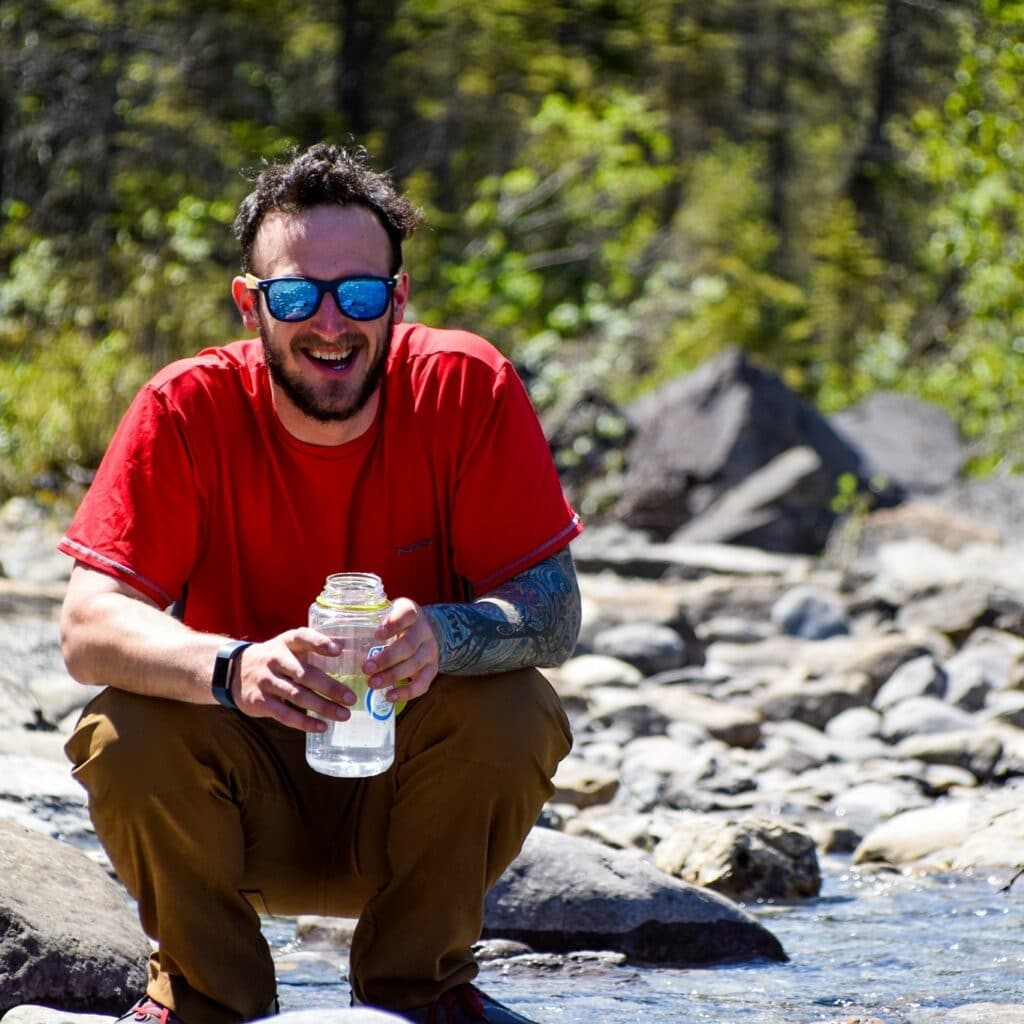 Smiling man wearing a red tshirt and sunglasses crouches next to a river holding waterbottle