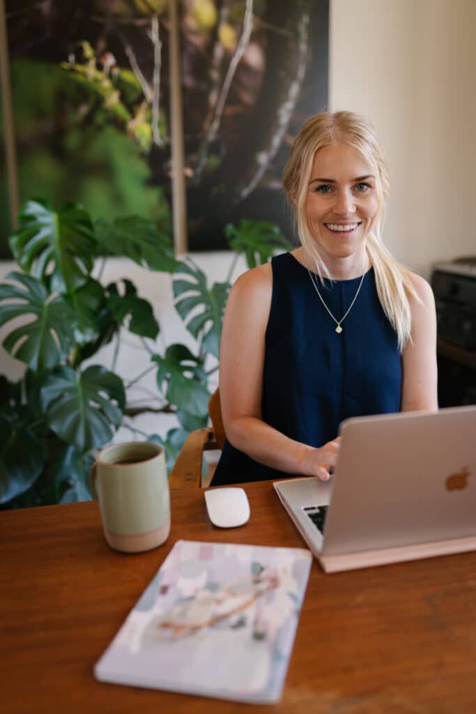 Smiling blonde woman wearing a black shirt and necklace sits at a laptop next to indoor plants