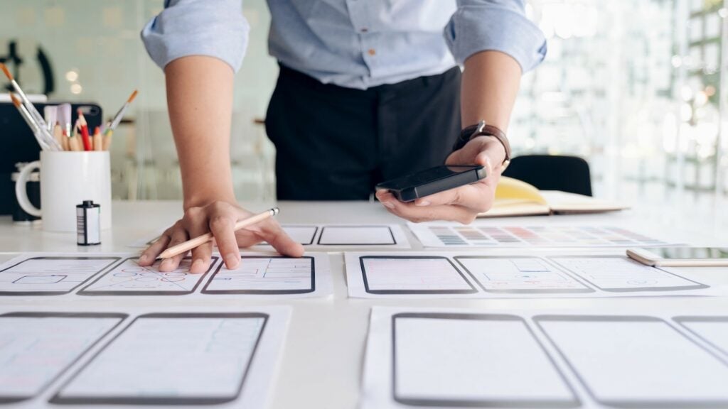 Man standing over a desk covered in papers holding a pencil and phone in each hand